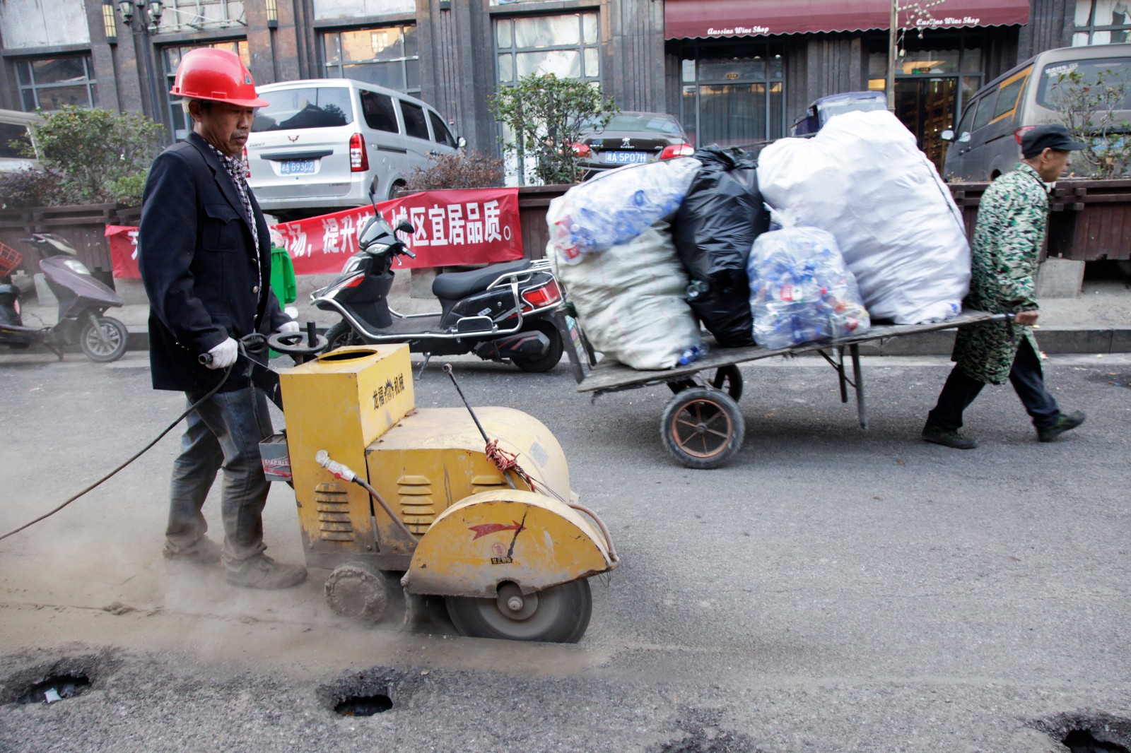 一名工人正在进行路面切割作业，旁边一名拉着一车废品的老人匆匆而过。