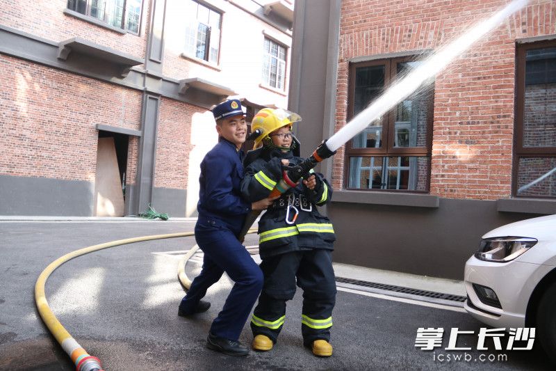 小朋友穿上灭火防护服，与消防员一起使用消防水枪“灭火”。